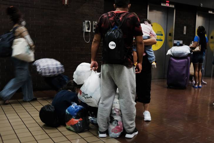 A man in grey sweatpants stands in the middle of the Port Authority Bus Terminal holding plastic bangs with his belongings as a woman holds a sleeping child.